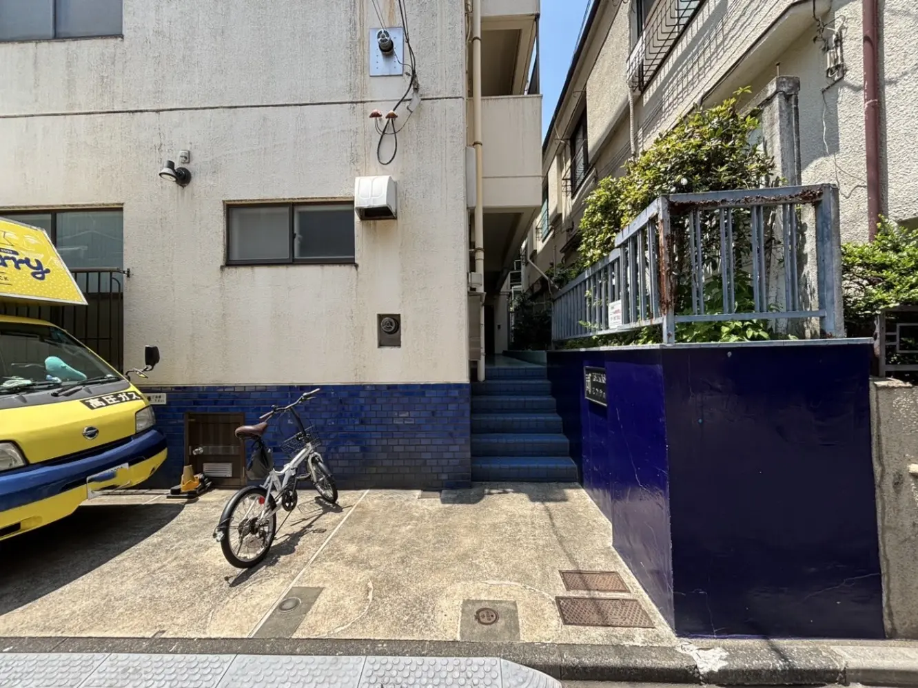 - 駐輪場に置かれた自転車と、青いタイルの階段が見えるアパートの外観、クリーム色の壁と青いアクセントのあるエリア