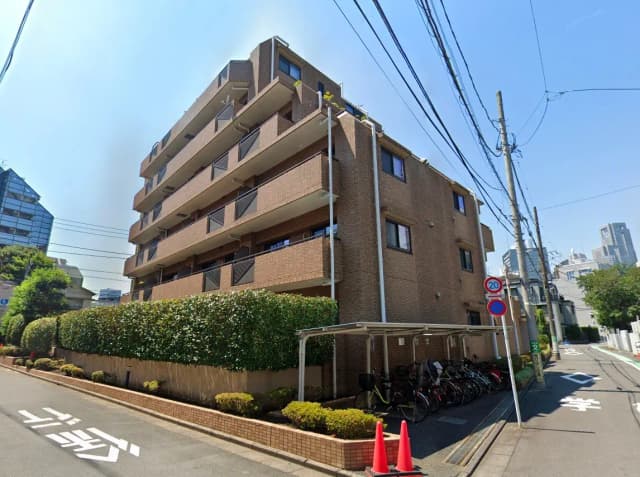 - 都市部にあるレンガ造りの5階建てアパートメント、周囲に緑の生け垣と自転車置き場、晴れた青空の下での外観写真