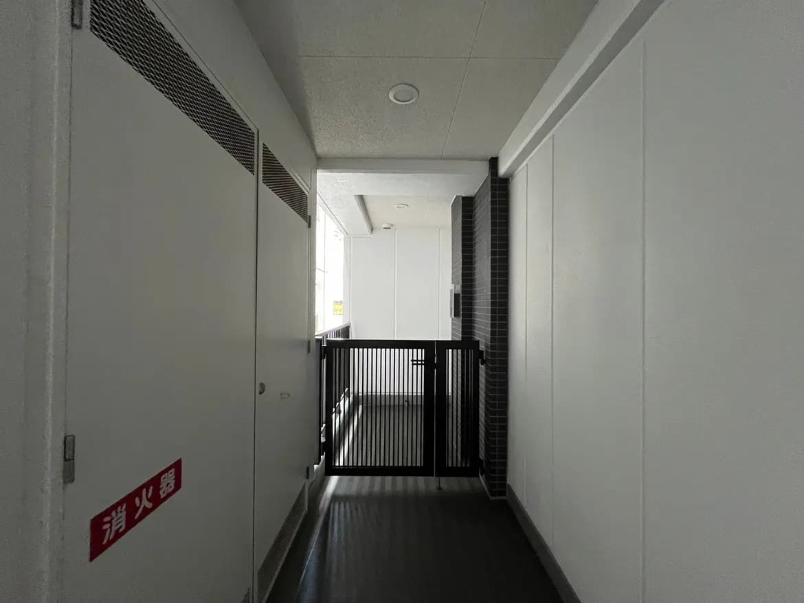 - 白い壁のある長い廊下、奥に開口部があり、手前には仕切り扉と消火器の標識が設置されたデザイン