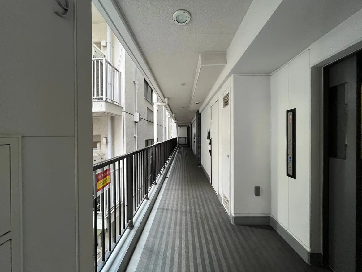 - 明るい色調のマンションの廊下、壁に沿ったバルコニーやエレベーターの近くに配置されたドアが並ぶ様子