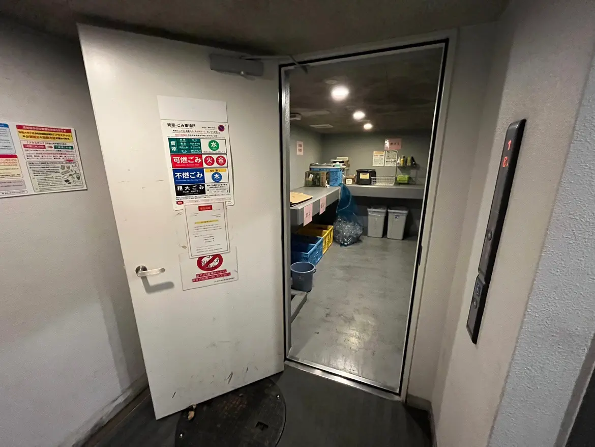- 清掃用具が整然と並ぶ倉庫の入り口、ゴミ分別の案内表示が貼られた白いドアと明かりのある室内が見える画像