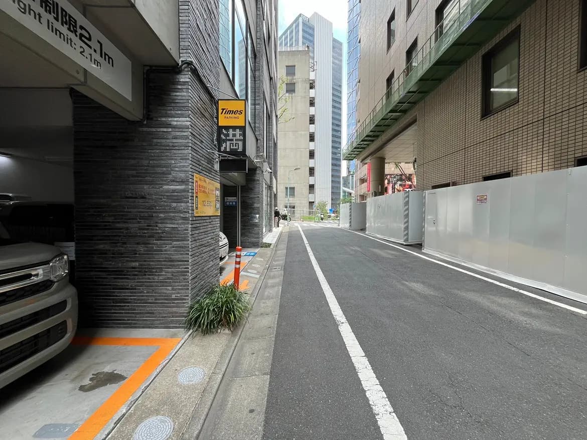 - 都市の駐車場入口、隣に見えるビルの外観と舗装された道路、横に並ぶパーキングサインと工事用のフェンスがある風景