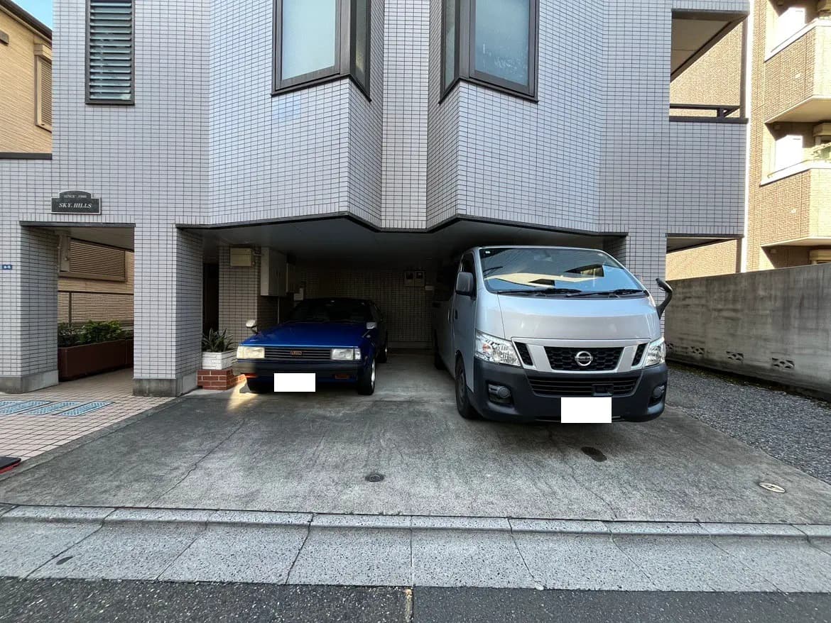 - 駐車スペースに停まっている青いセダンとシルバーのバン、周囲のタイル張りの建物と植栽の様子