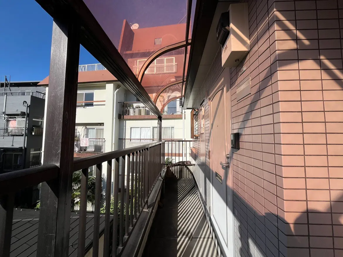 - 日差しが差し込むアパートの廊下、手すり付きのバルコニーと隣接する部屋の窓が見える風景