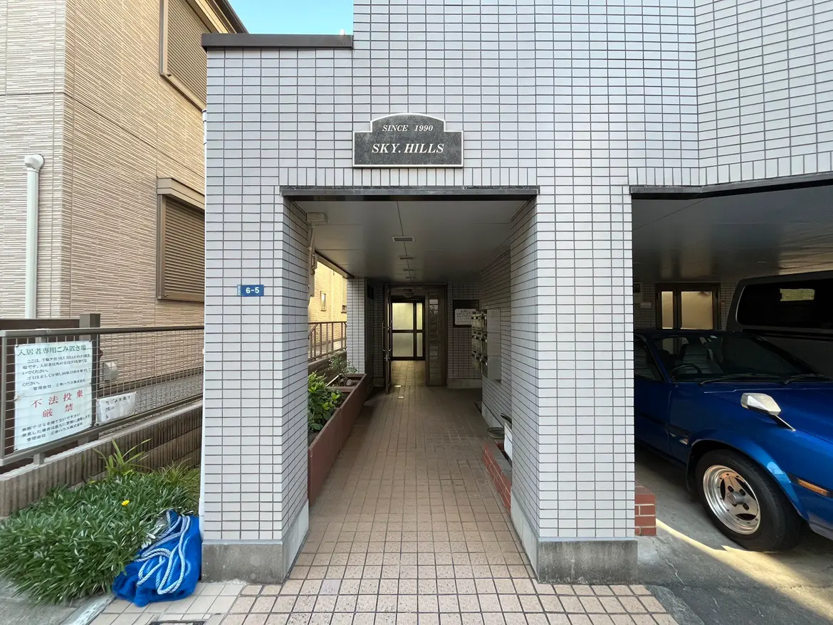 - 都市型マンションのエントランス、タイル張りの通路と駐車スペース、周囲に植栽があるエリア