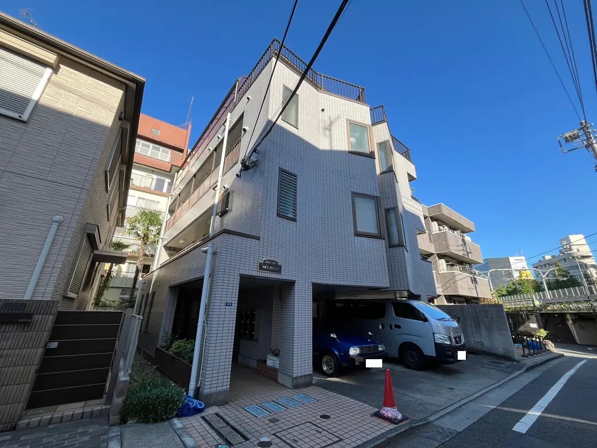- 都市部に位置する白タイルの外観を持つ中層マンション、青空を背景にした建物の立体感と周囲の景観