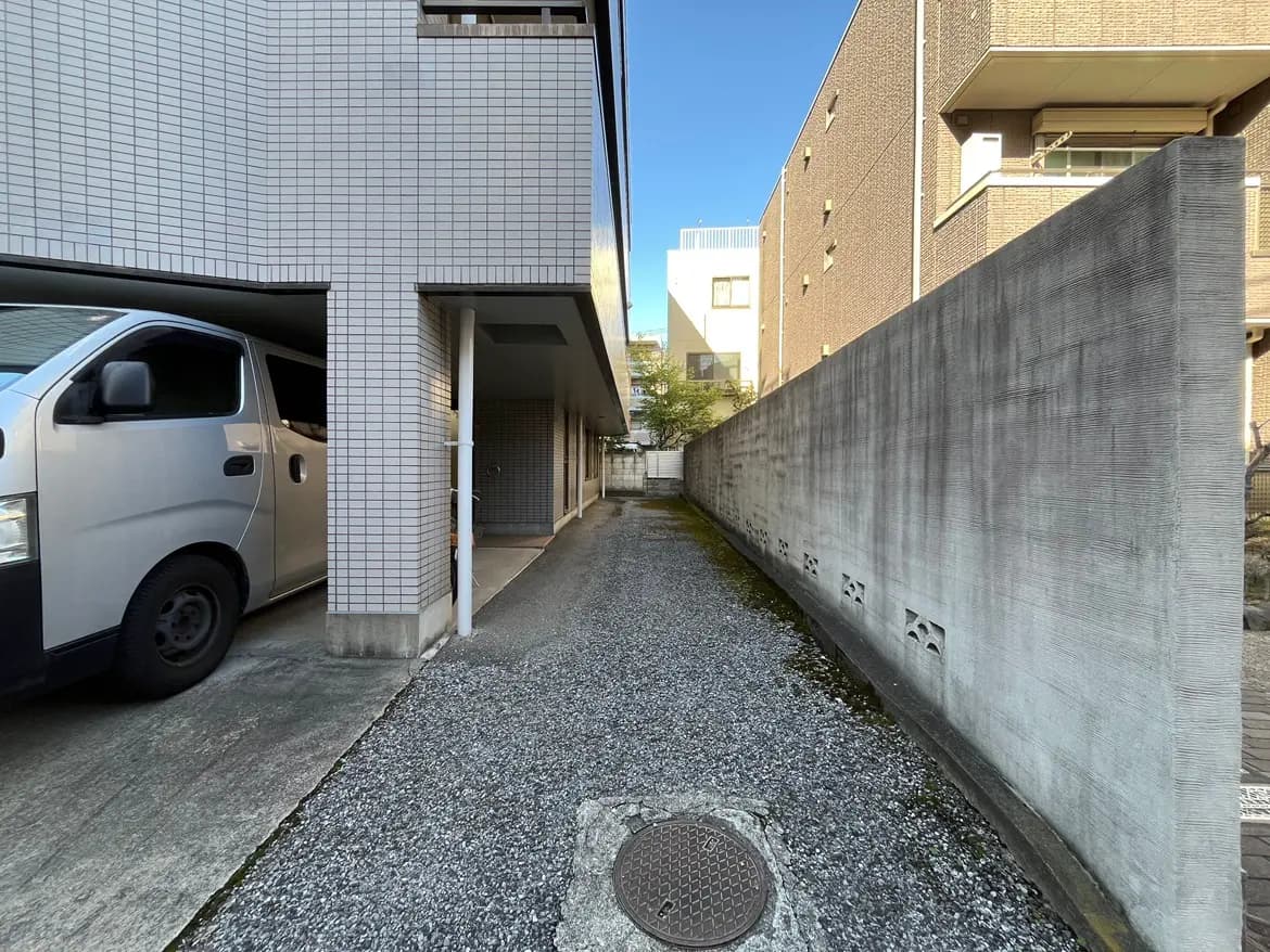 - 駐車スペースと住宅の間の細長い通路、左側に白いバンと右側にコンクリートの壁が見える風景