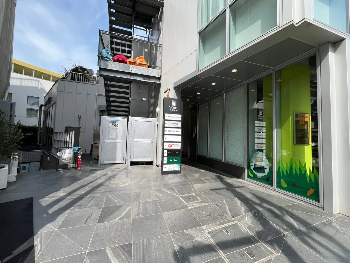 - モダンなビルの入口、ガラス張りのエントランスと石材の床、隣接する階段と看板がある風景