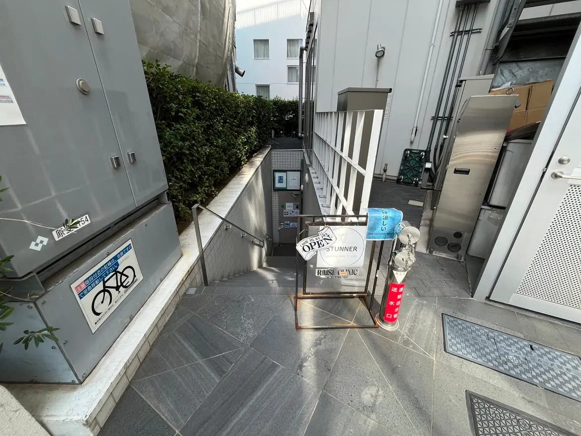 - 階段下にあるカフェの入り口、看板が「OPEN」と表示され、周囲には緑の植物と洗練されたデザインの壁が目を引く街の風景