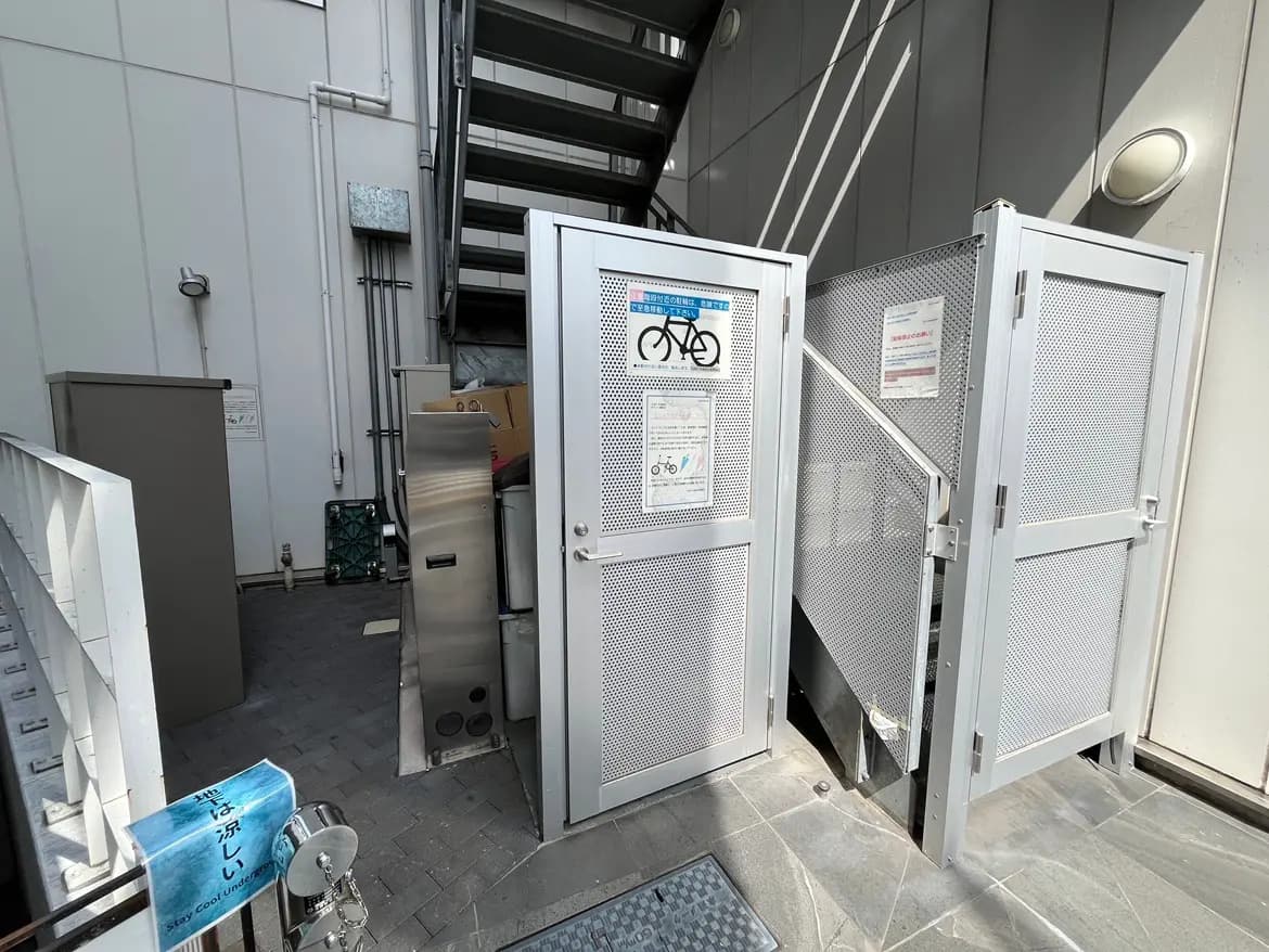 - 自転車通行禁止サインが掲示された、金属製のフェンスに囲まれた外の自転車置き場、階段と周囲の構造物が写る風景