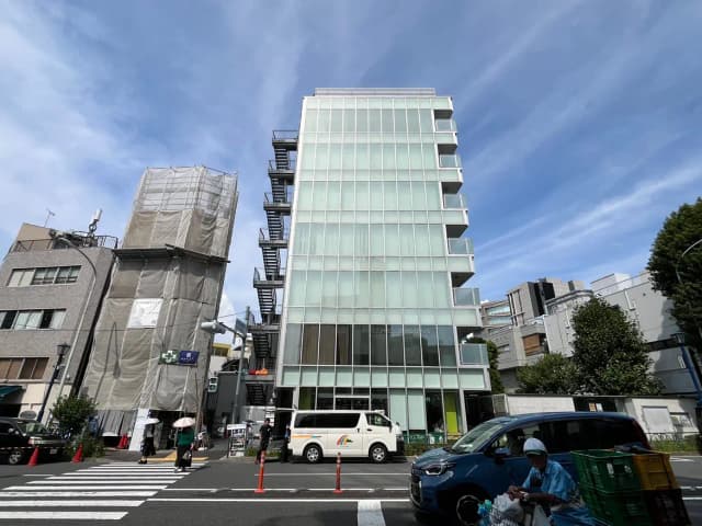 モダンなガラス張りのビルが並ぶ都市の風景、青空と白い雲が広がり、横断歩道を渡る人々や通行する車が写るシーン