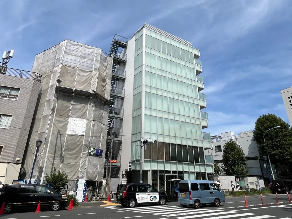 - 施工中の現代的なビルの外観、ガラス面と工事用ネットがかけられた部分、青空と道路の様子