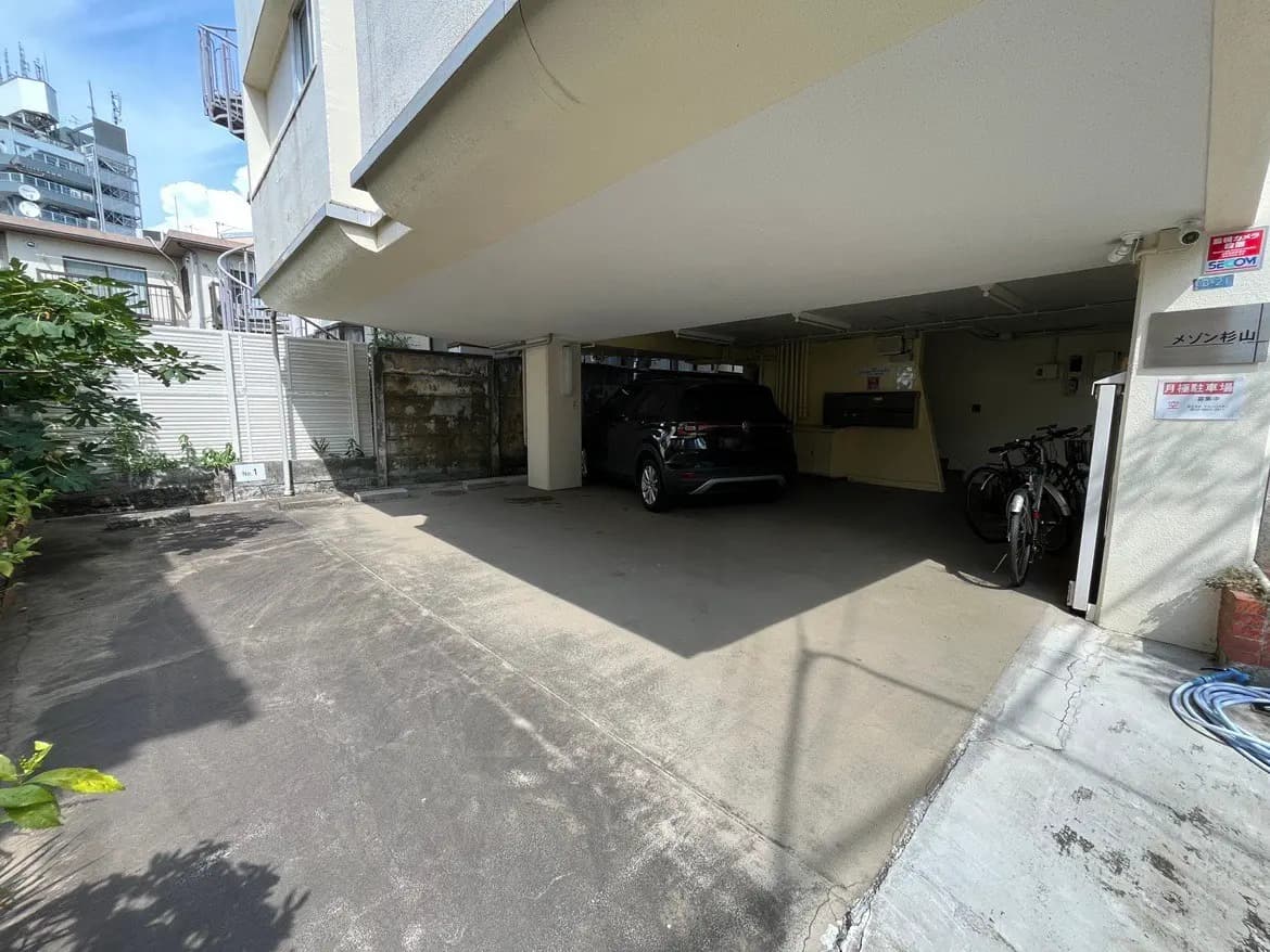 - 駐車場の屋根付きスペース、黒い車と自転車が置かれた清潔感ある外観、周囲には緑の植物が見える