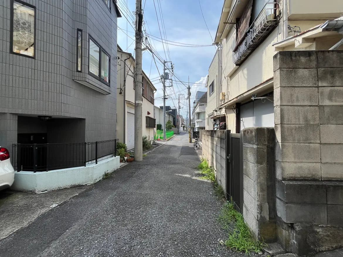 - 街中の住宅街の一角、左右に住宅が並ぶ細い路地、青空と電線が見える風景