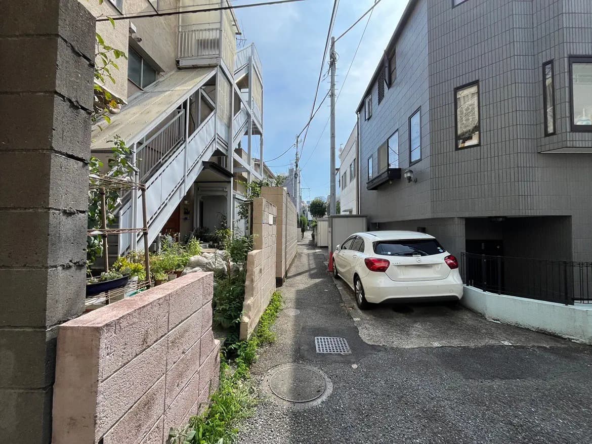 - 狭い路地に面した住宅街の風景、階段状の集合住宅と白い車が停まる駐車スペース、緑の植物が点在する情景