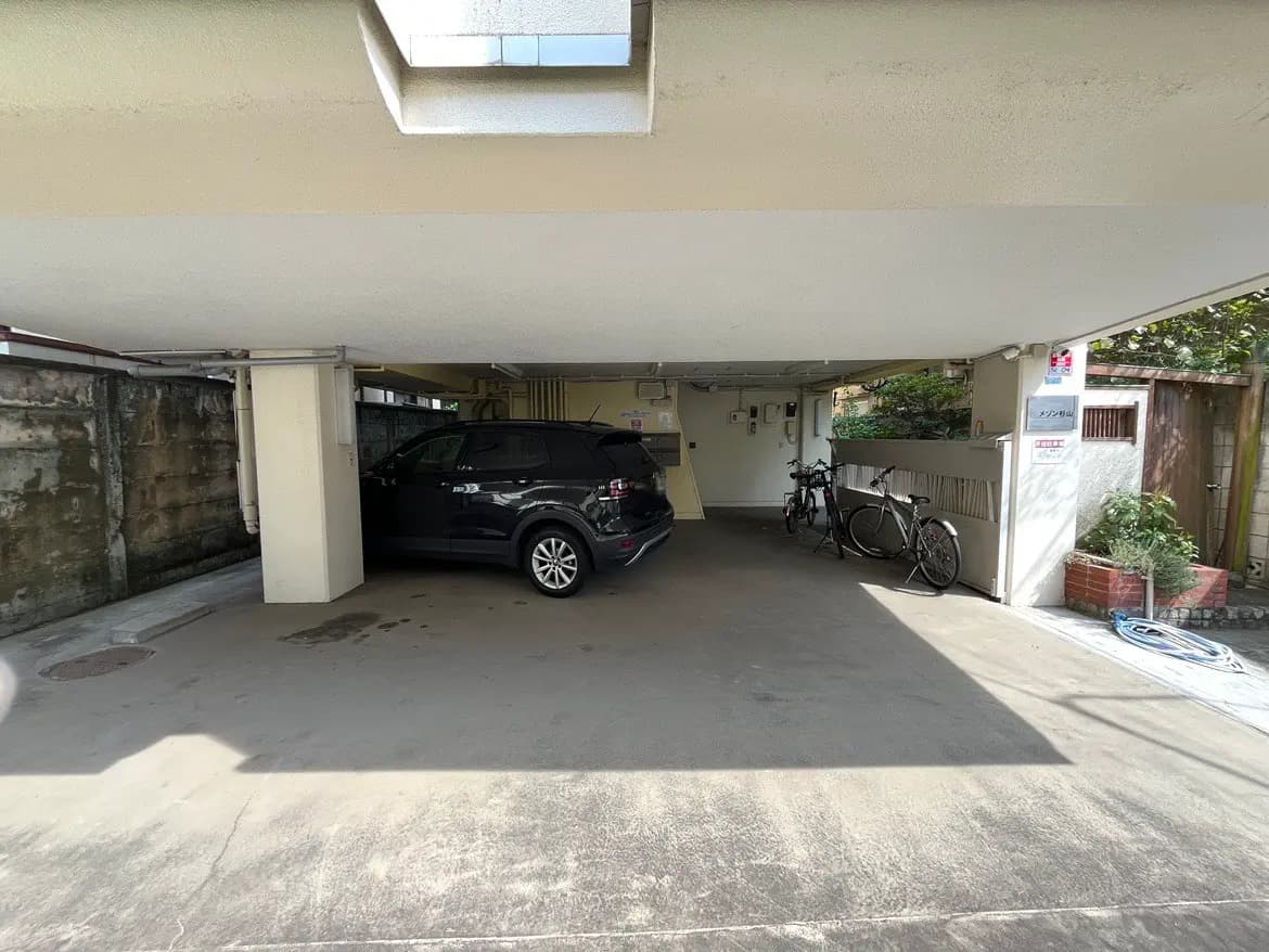 - 屋根付きの駐車スペースに停まっている黒いSUVと自転車、周囲に緑豊かな植物がある景観