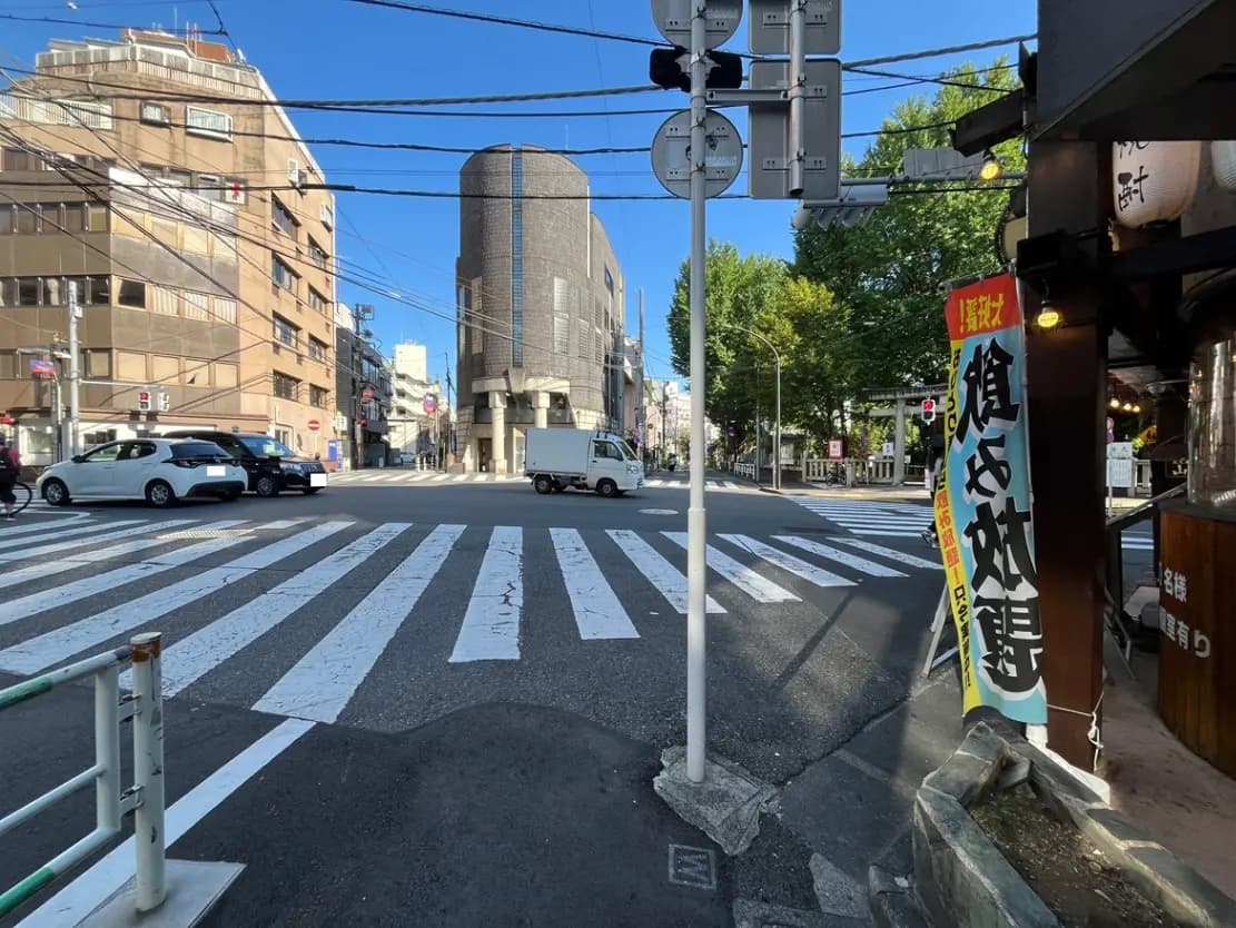 - 都市の交差点、白い横断歩道と青空、周囲のビルや街路樹が見える風景、右側には飲食店の看板があるシーン