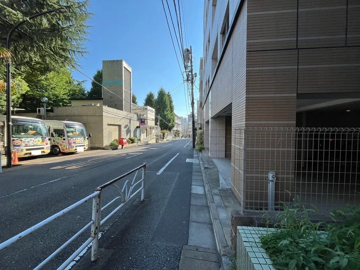 - 日差しの差し込む静かな街道、コンクリートの建物と緑の木々、並んだバスと歩道の風景