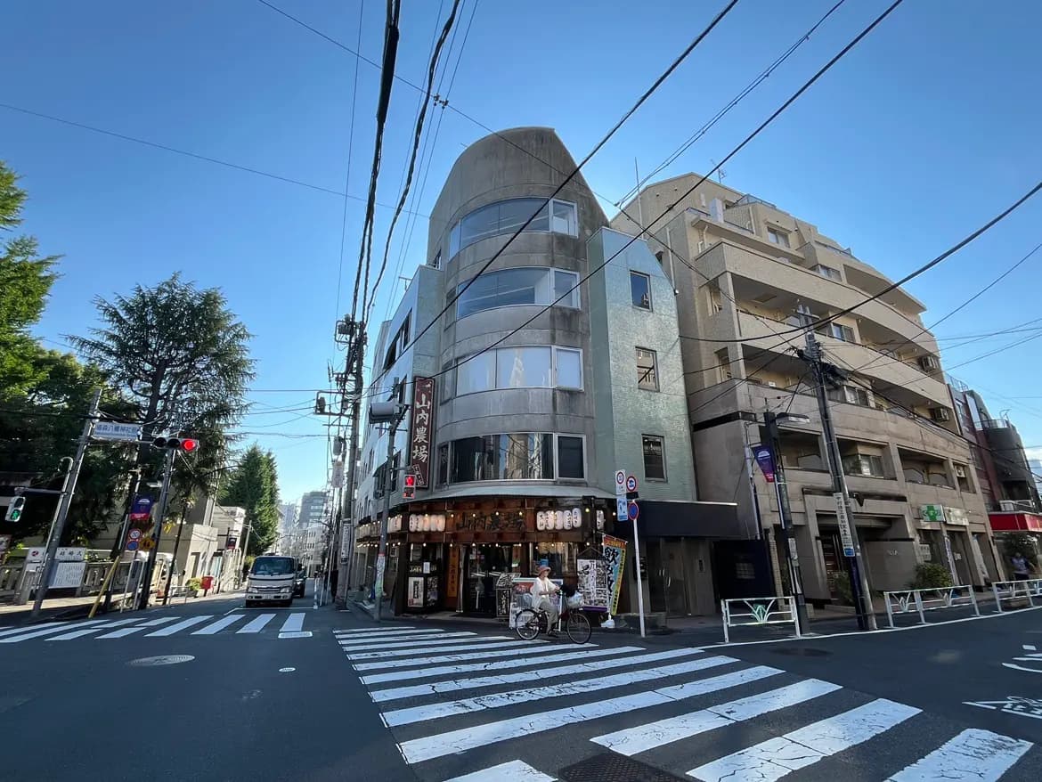 - 都市の交差点に位置する現代的な建物、手前には横断歩道と信号機、青空が広がる風景