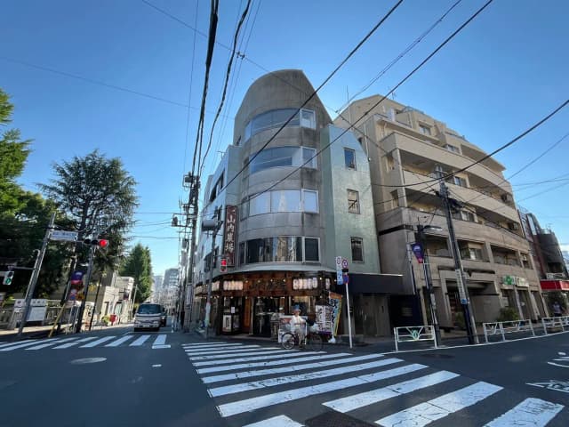 - 都市の交差点に位置する現代的な建物、手前には横断歩道と信号機、青空が広がる風景