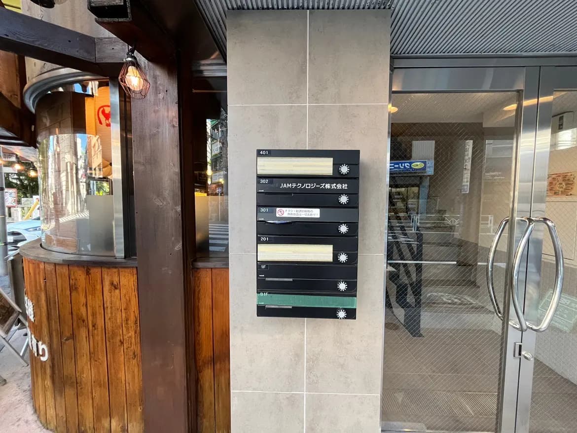 - オフィスビルの入り口に設置された複数の表札が並ぶ掲示板、周囲に木製の壁とガラス張りのインテリアが見える風景
