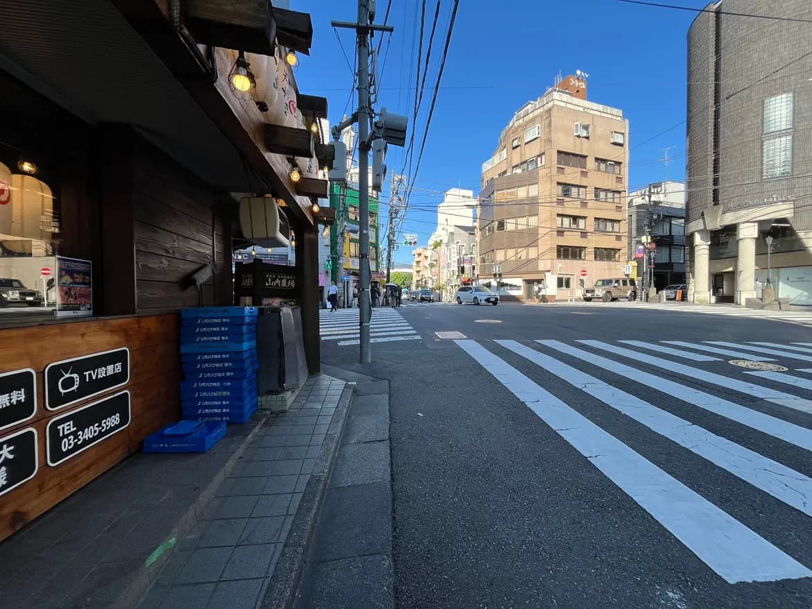 - 交差点に面した飲食店の外観、横には青い箱が積まれ、周囲にはビルと青空が広がる街の風景