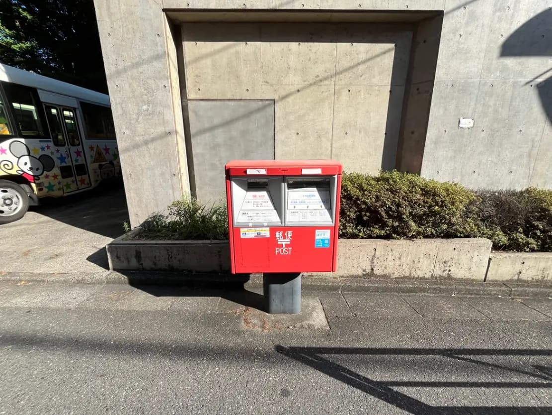 - コンクリート壁の前に設置された赤い郵便局の投函箱、周囲には緑の植栽と通り道が見えるシーン