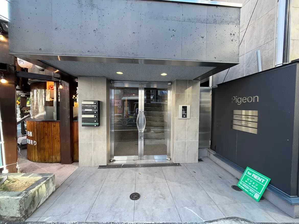 - モダンなデザインの集合住宅のエントランス、ガラスの自動ドアと郵便受け、近くには賃貸看板が立てられた外観