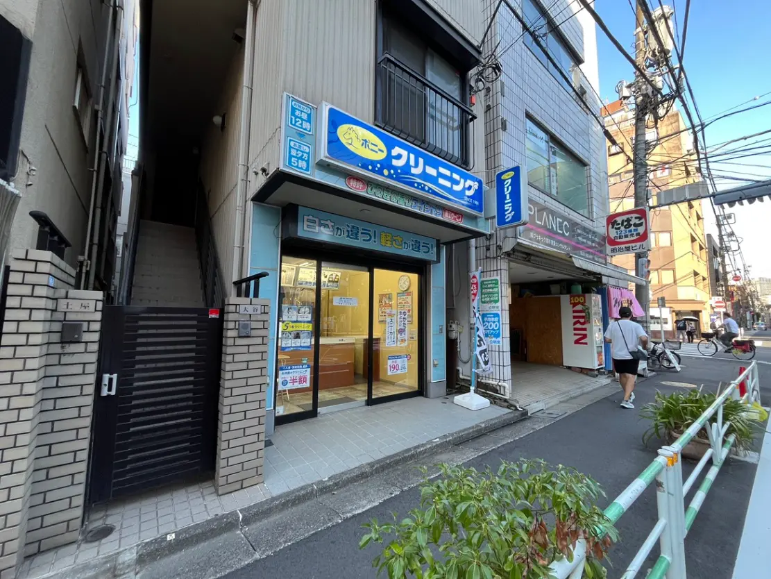 - 街角に位置するクリーニング店の外観、明るい看板と隣接する階段、周囲には通行人や自転車が見える風景
