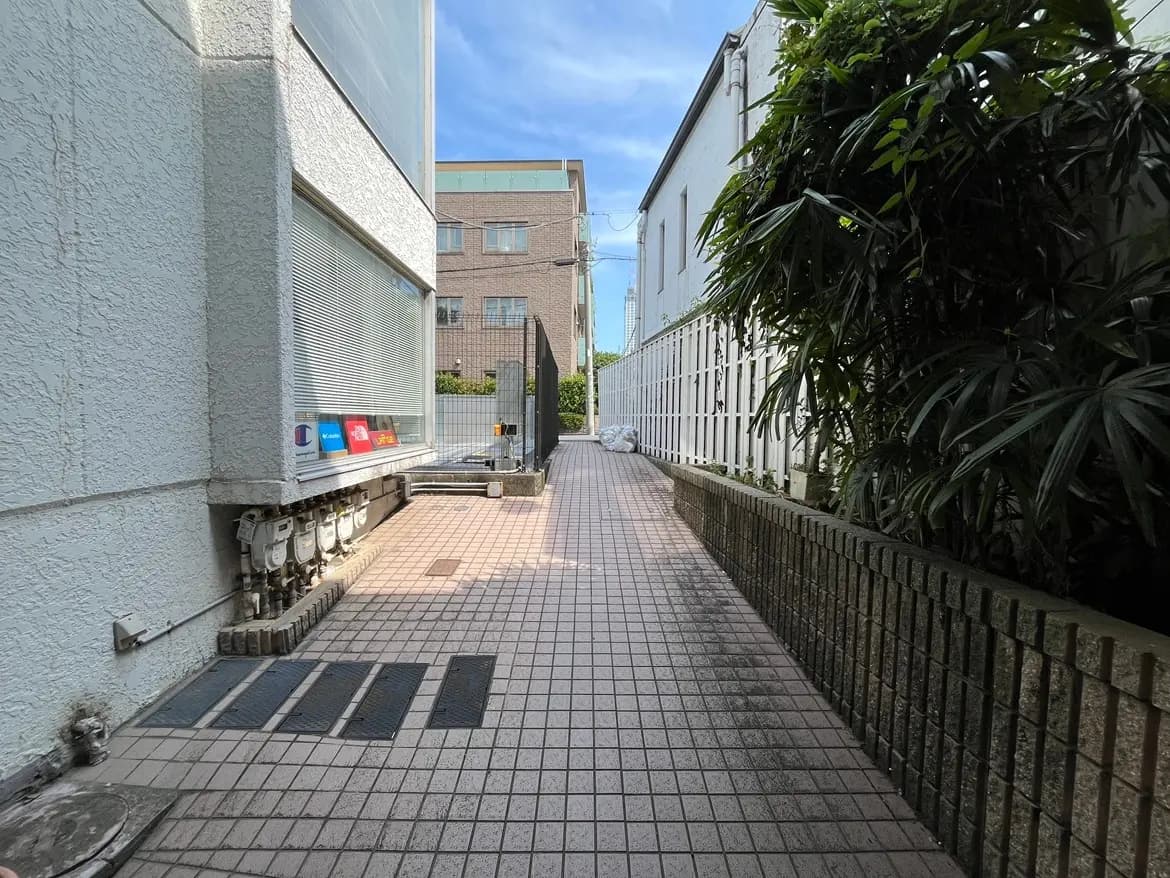 - 建物の横にある舗装された通路、緑豊かな植物と白いフェンスが囲む風景、明るい青空が広がるシーン