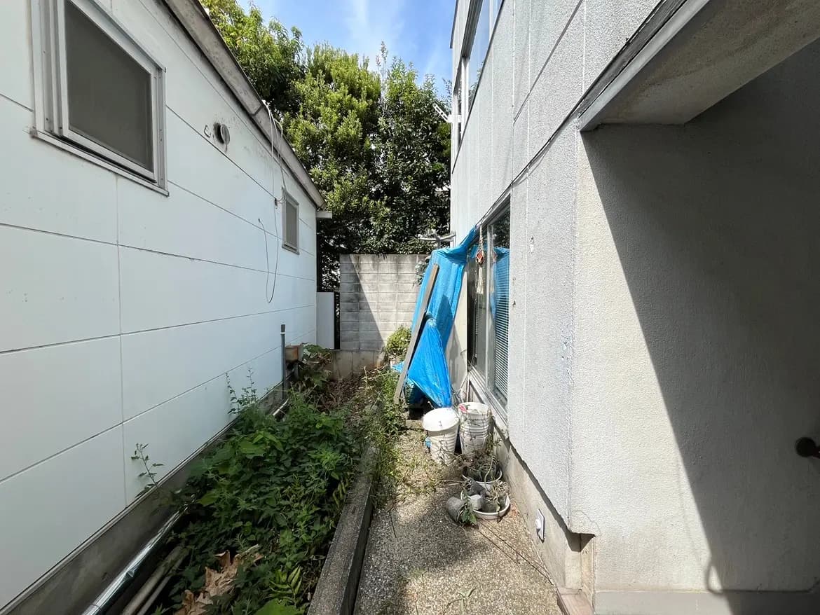 - 隣接する2つの建物の間のスペース、青いシートがかぶせられたエリアと雑草の生えた庭の様子