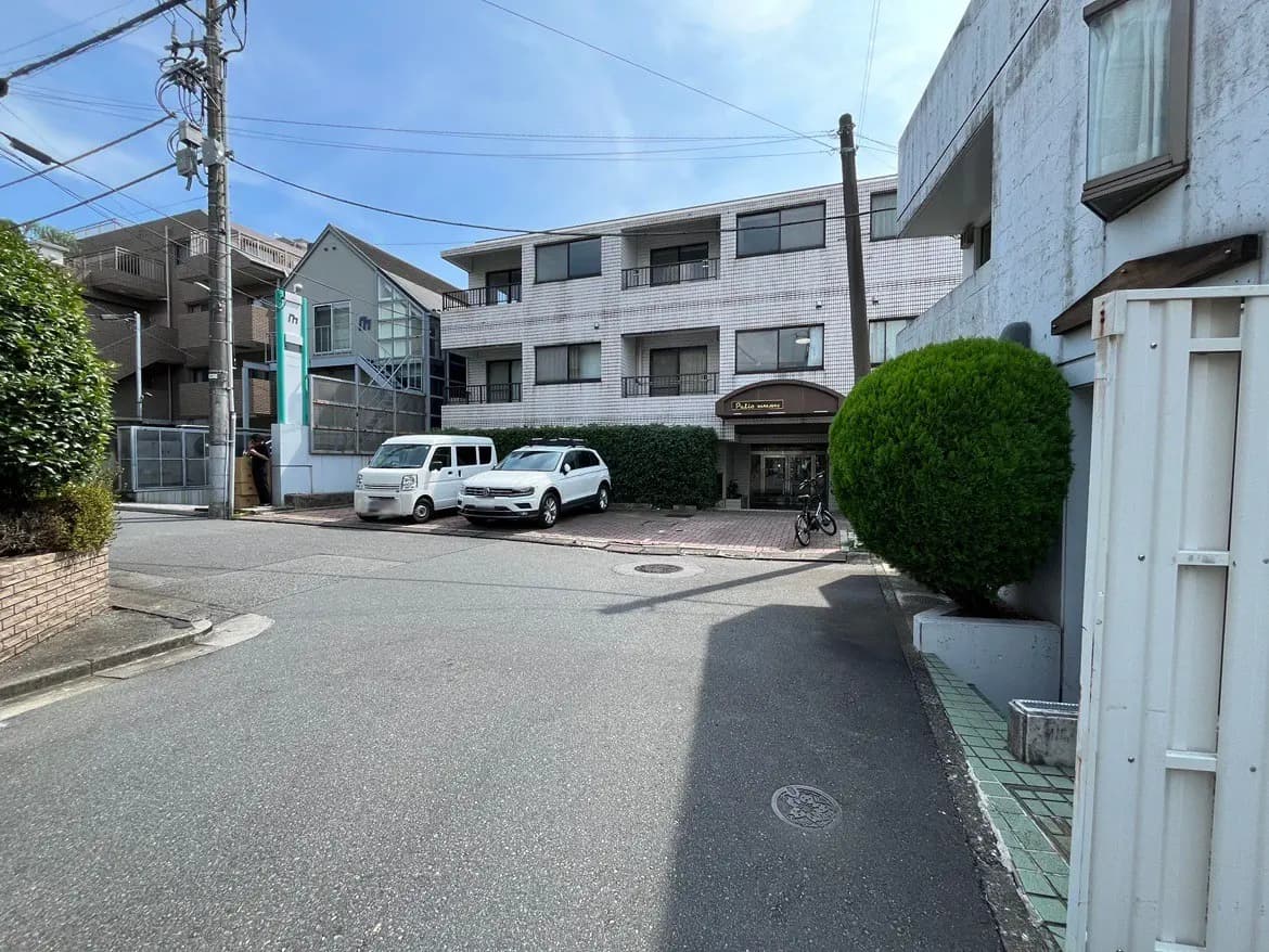 - 住宅街の角に位置するマンションの外観、白い車と緑の庭木が並ぶ風景、青空が広がる日中のシーン