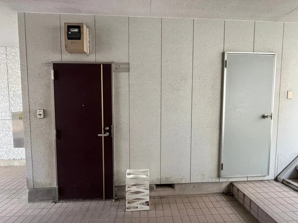 - シンプルな廊下の壁面に設置されたダークカラーのドアと小さな灰色のドア、隣に白い折りたたみ式の椅子が置かれた空間