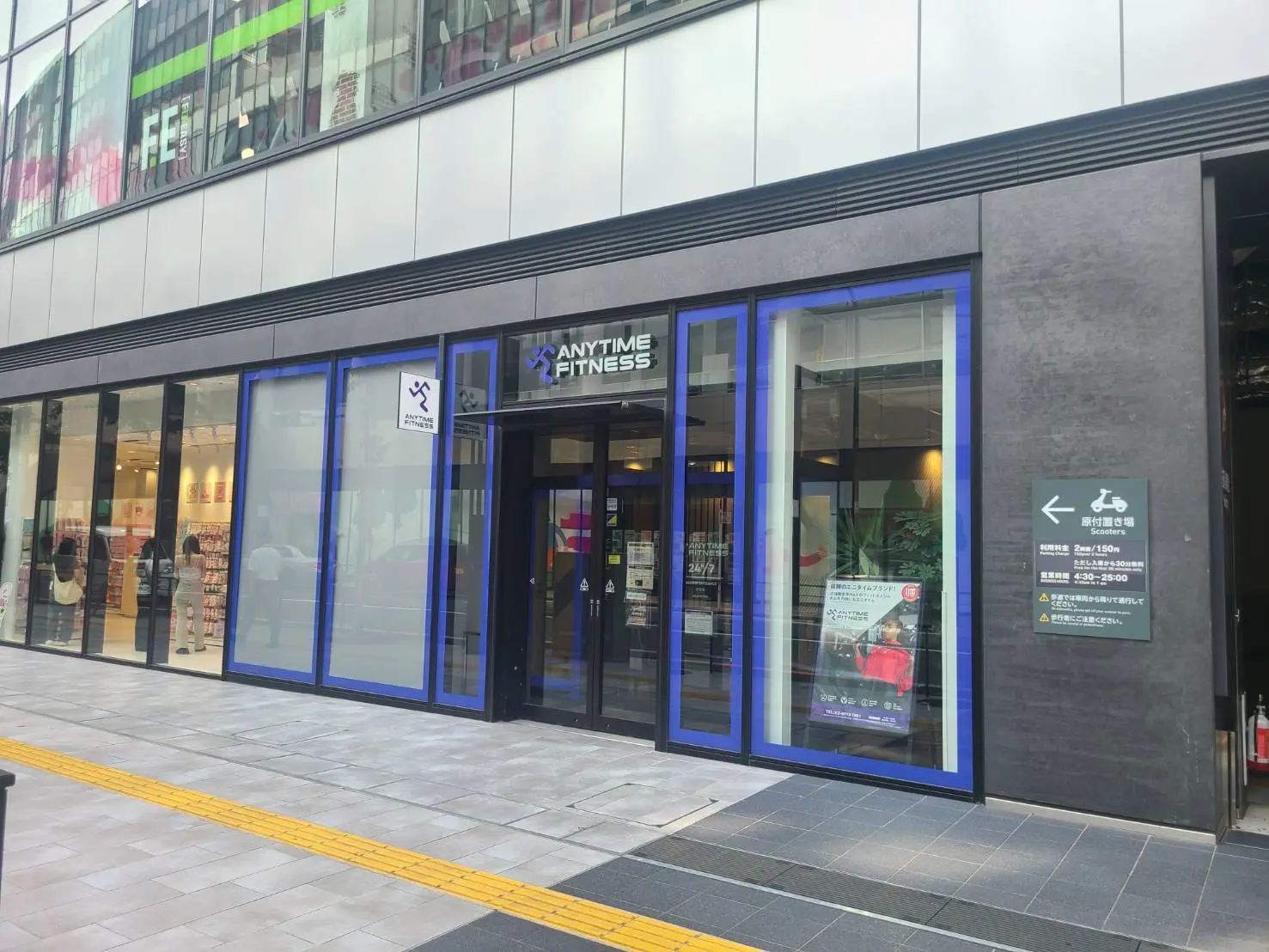 - 24時間営業のフィットネスジム「ANYTIME FITNESS」の外観、明るいガラス張りの店舗と入り口、近くのショップが見える景観