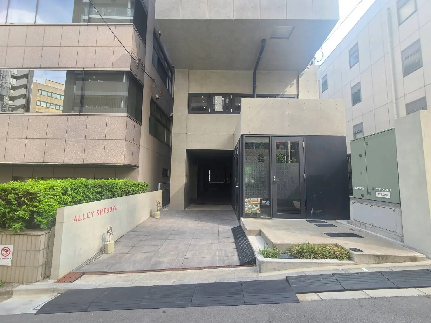 - シンプルでモダンな外観の建物入り口、コンクリートの壁に囲まれた通路とALLEY SHIBUYAのサイン、緑の植栽が施されたエントランス