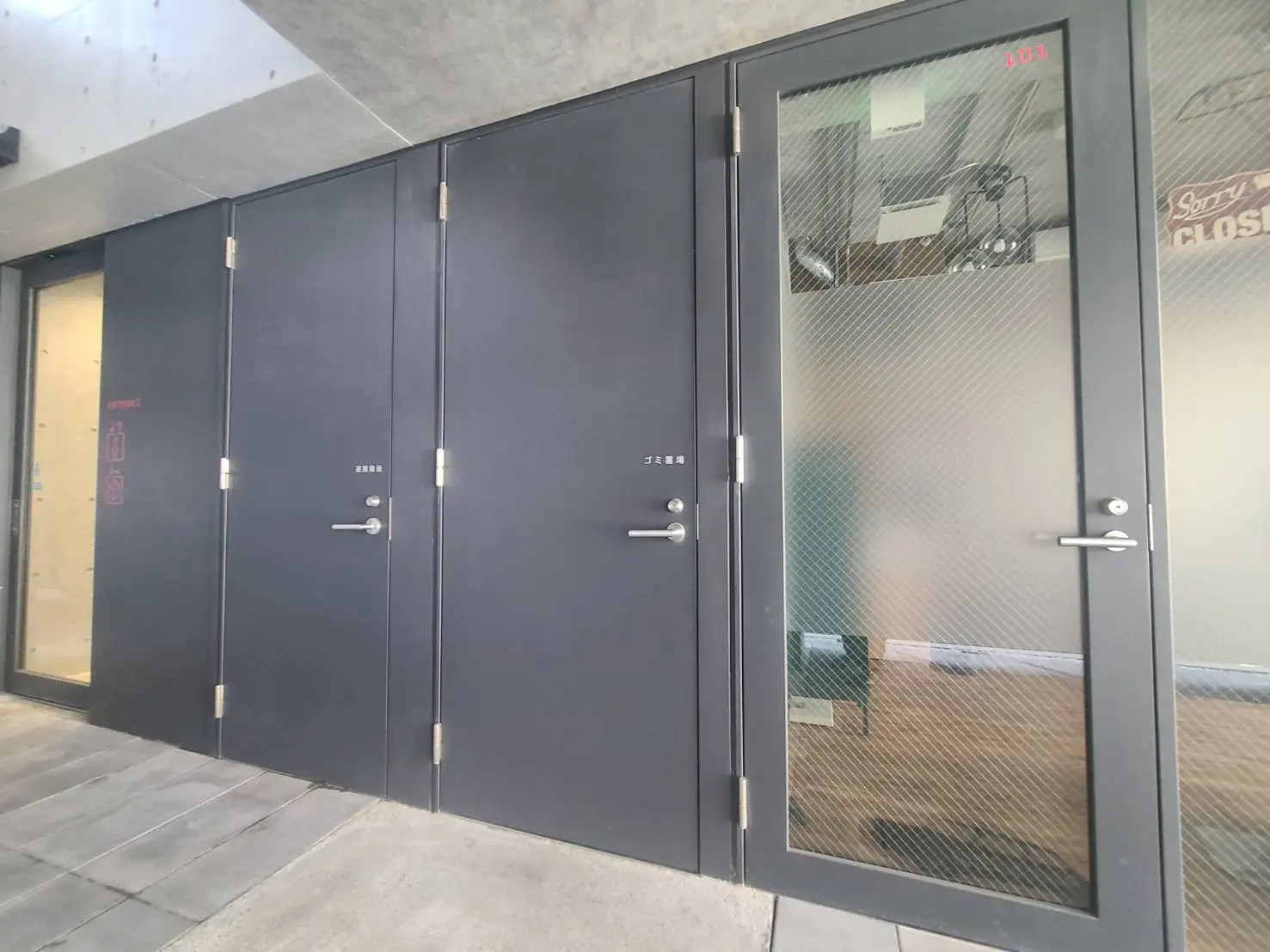 モダンなデザインの建物入口、スモークガラスと金属製のドアが並ぶ外観、周囲のコンクリートと調和したシンプルなスタイル