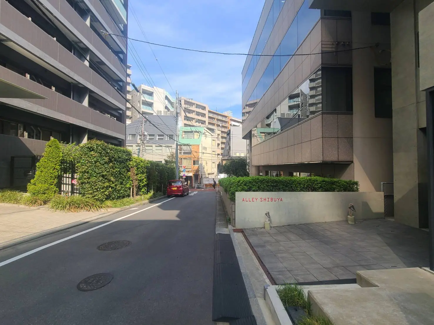- 渋谷の静かな路地、両側に高層ビルと緑豊かな植栽が並ぶ風景、赤い車が通りを走る様子
