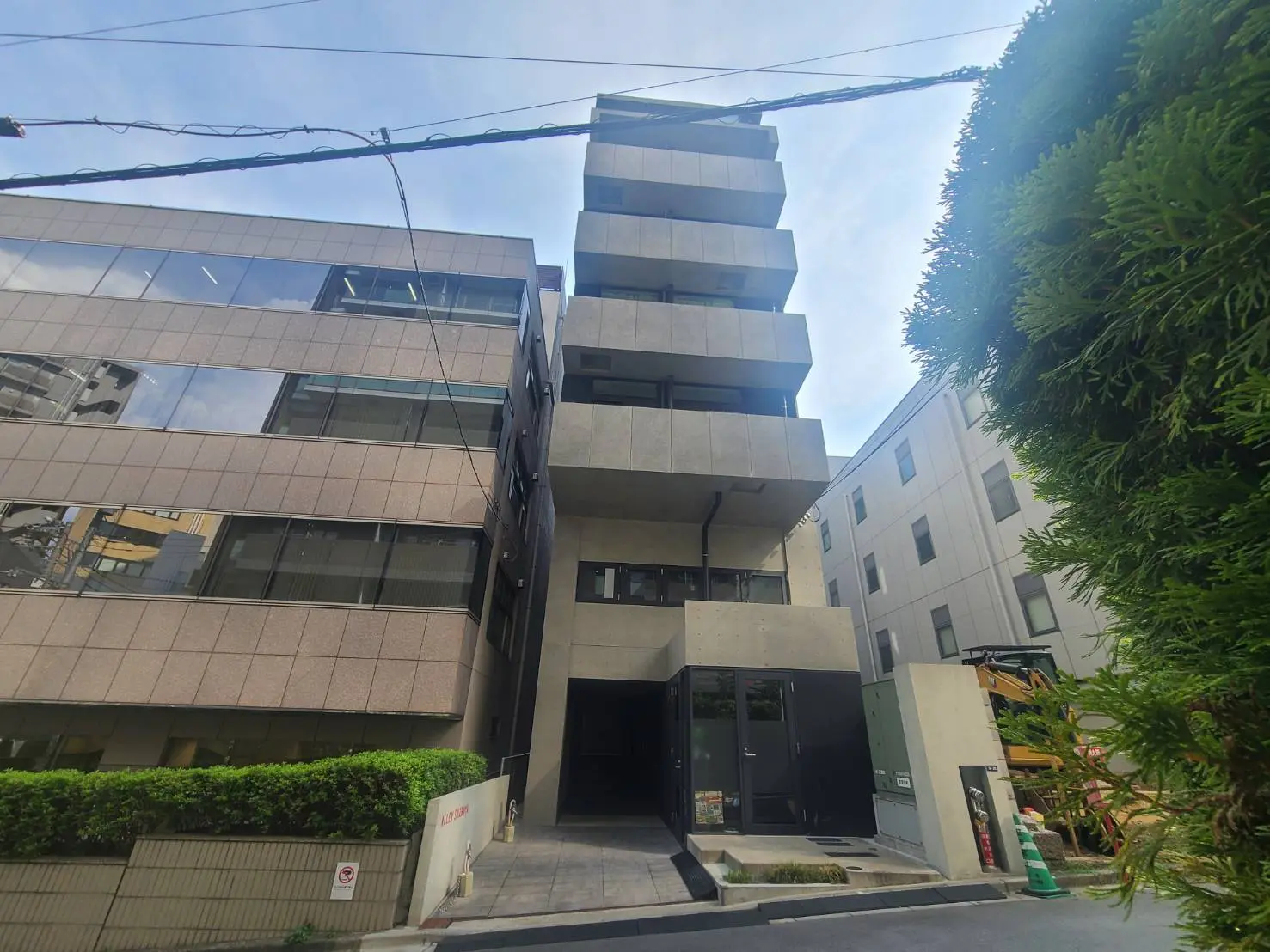 - モダンな外観のビル、グレーのタイル張りと大きな窓、エントランス前の緑豊かな植栽が特徴的な都市の風景