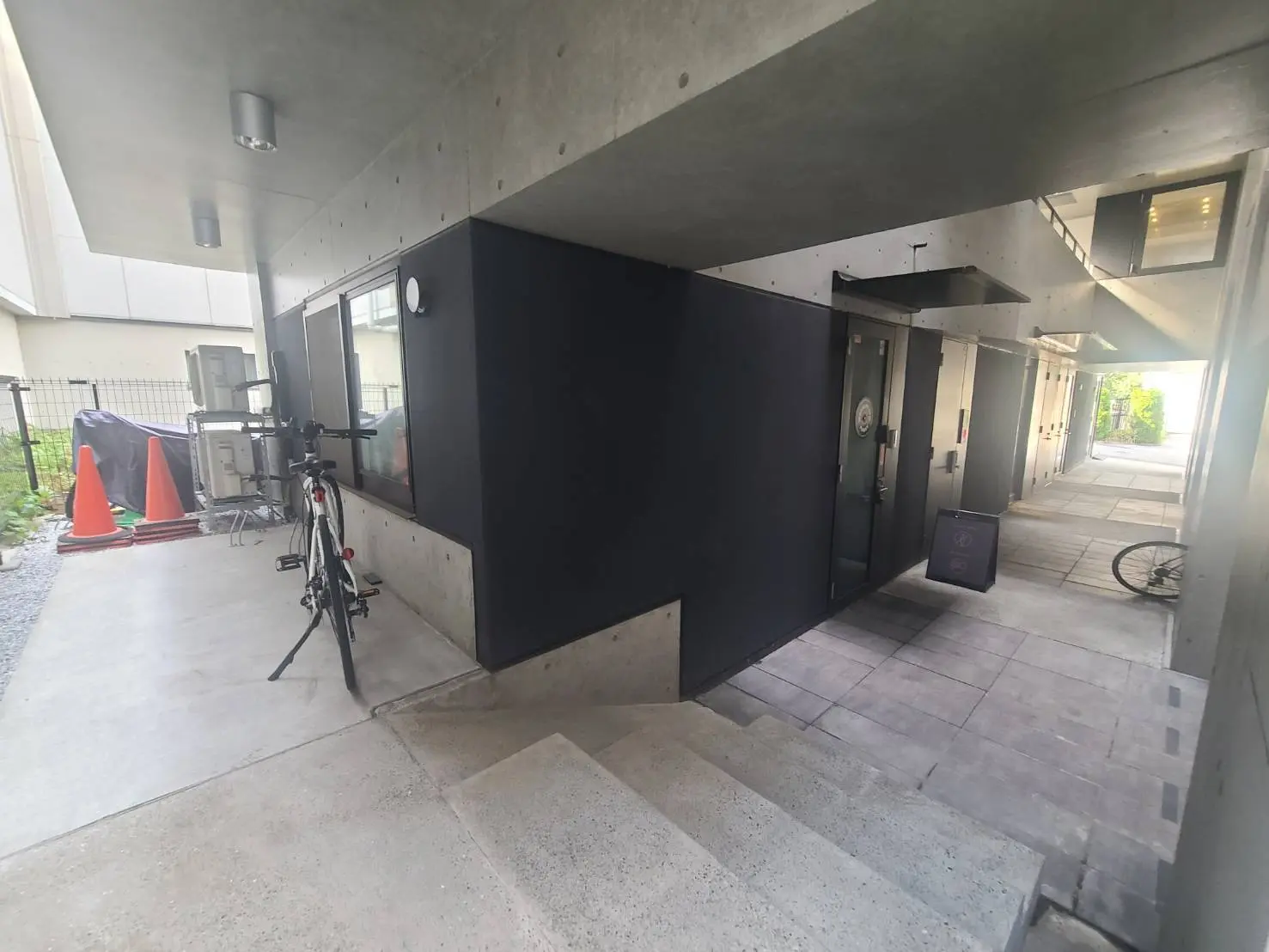 - コンクリート製の外観を持つモダンな建物のエントランス、左側に自転車とオレンジ色のコーンが並ぶ風景