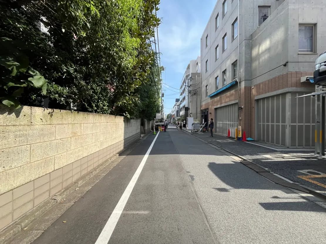 - 明るい青空の下、緑豊かな街路沿いの風景、コンクリート製の壁と商業ビルが並ぶ静かな通りの一角