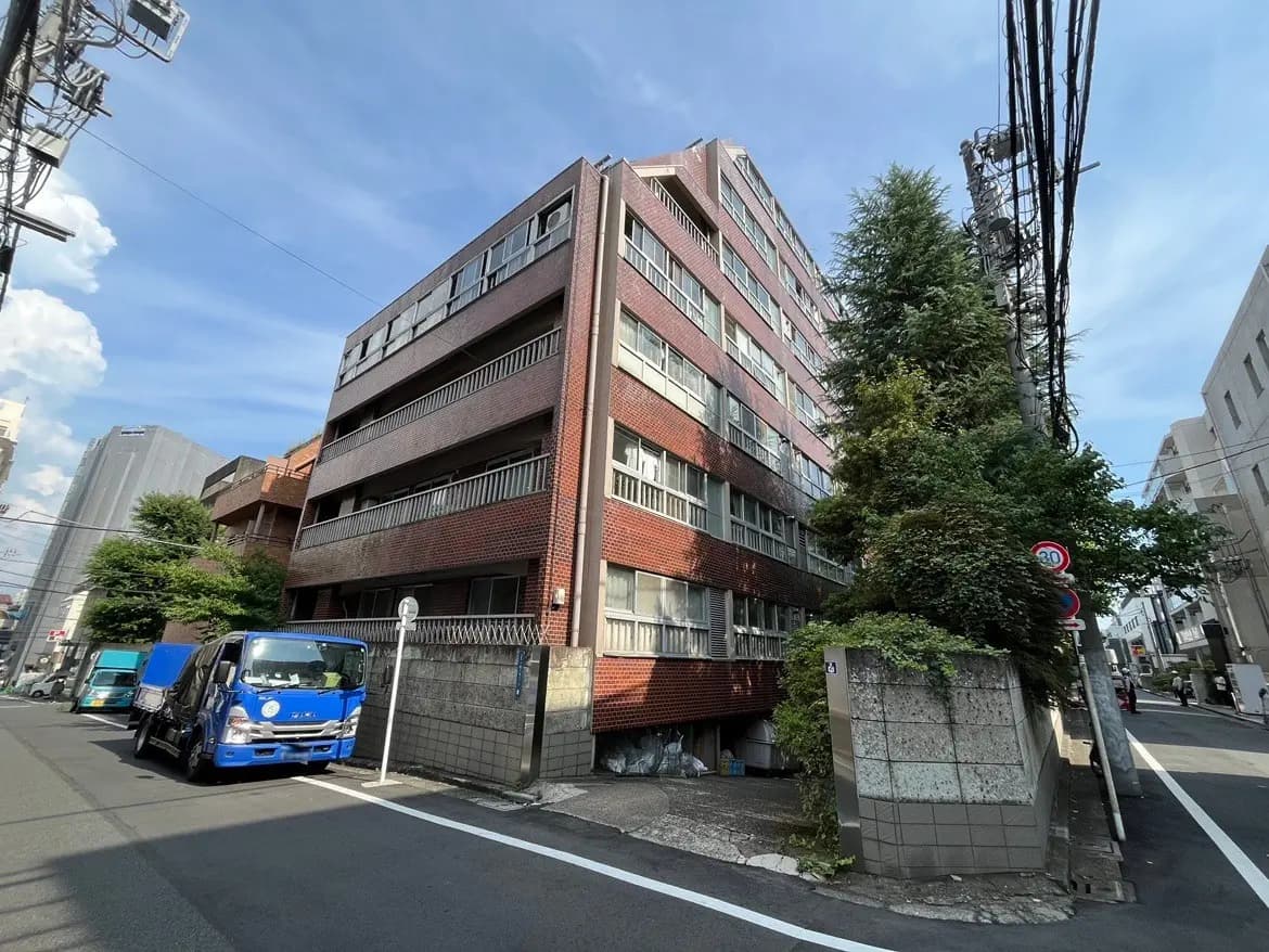 - 駐車場の前に立つレンガ造りの中層マンション、緑豊かな庭木と青空の下、周囲には商業ビルが見える街並み