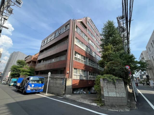 - 駐車場の前に立つレンガ造りの中層マンション、緑豊かな庭木と青空の下、周囲には商業ビルが見える街並み