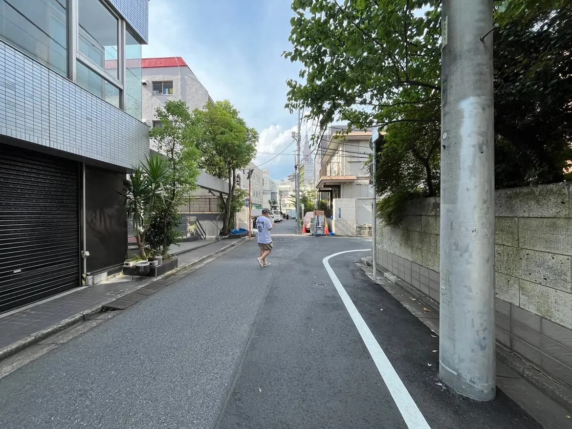 - 都市の住宅地における細い道路、左側に黒いシャッターの商業ビル、右側には緑の木々とブロック塀が立ち並ぶ風景、歩道を歩く人の姿