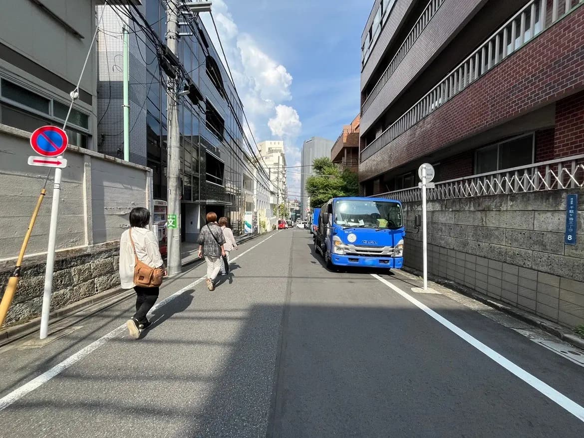 - 都会の街並みを歩く人々、青いトラックが停まる路地、近代的な建物と青空が広がる景色