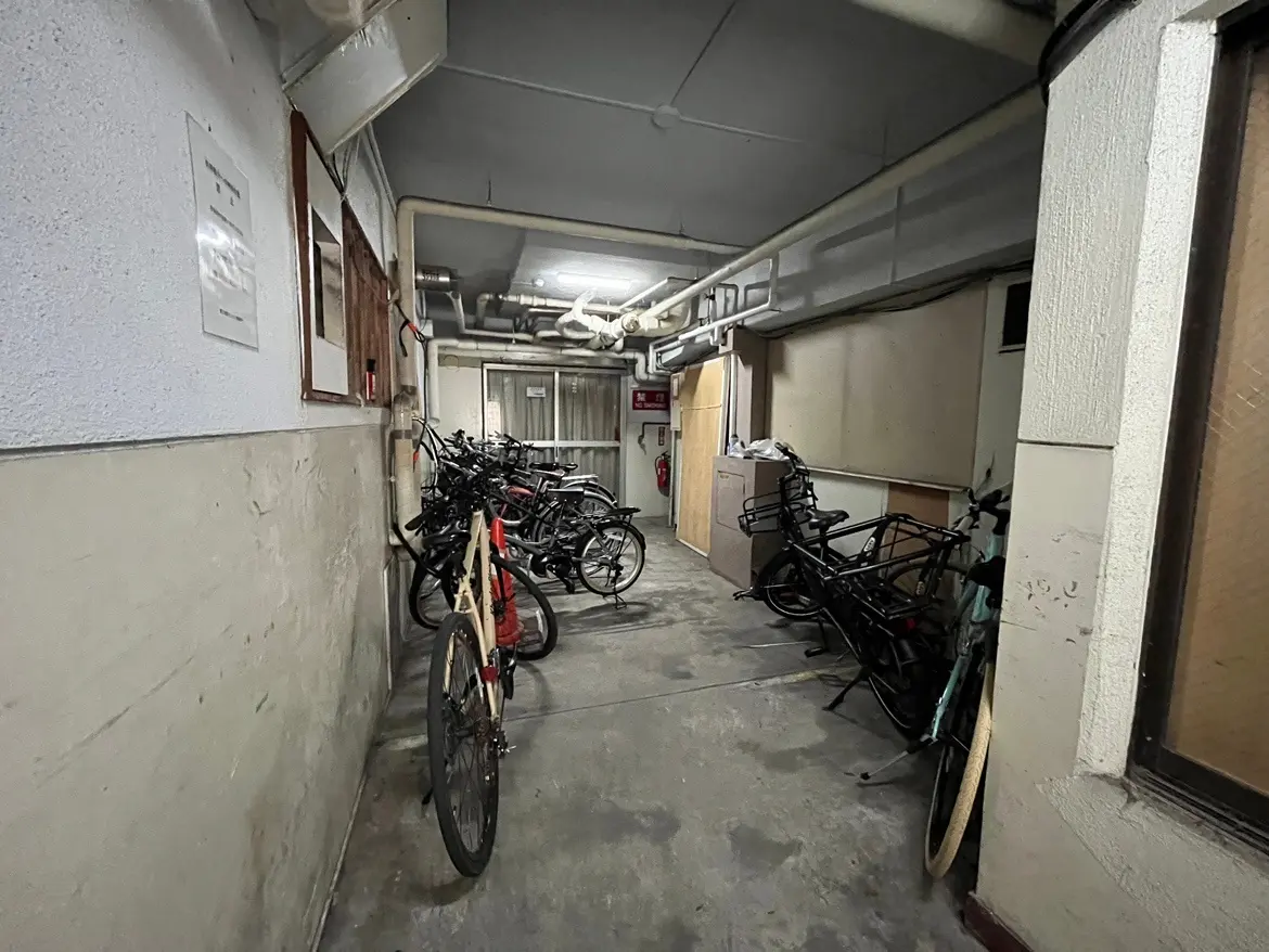 狭い通路に多くの自転車が停められた状況、壁は無機質な素材で構成され、天井には配管が見える内部のシーン