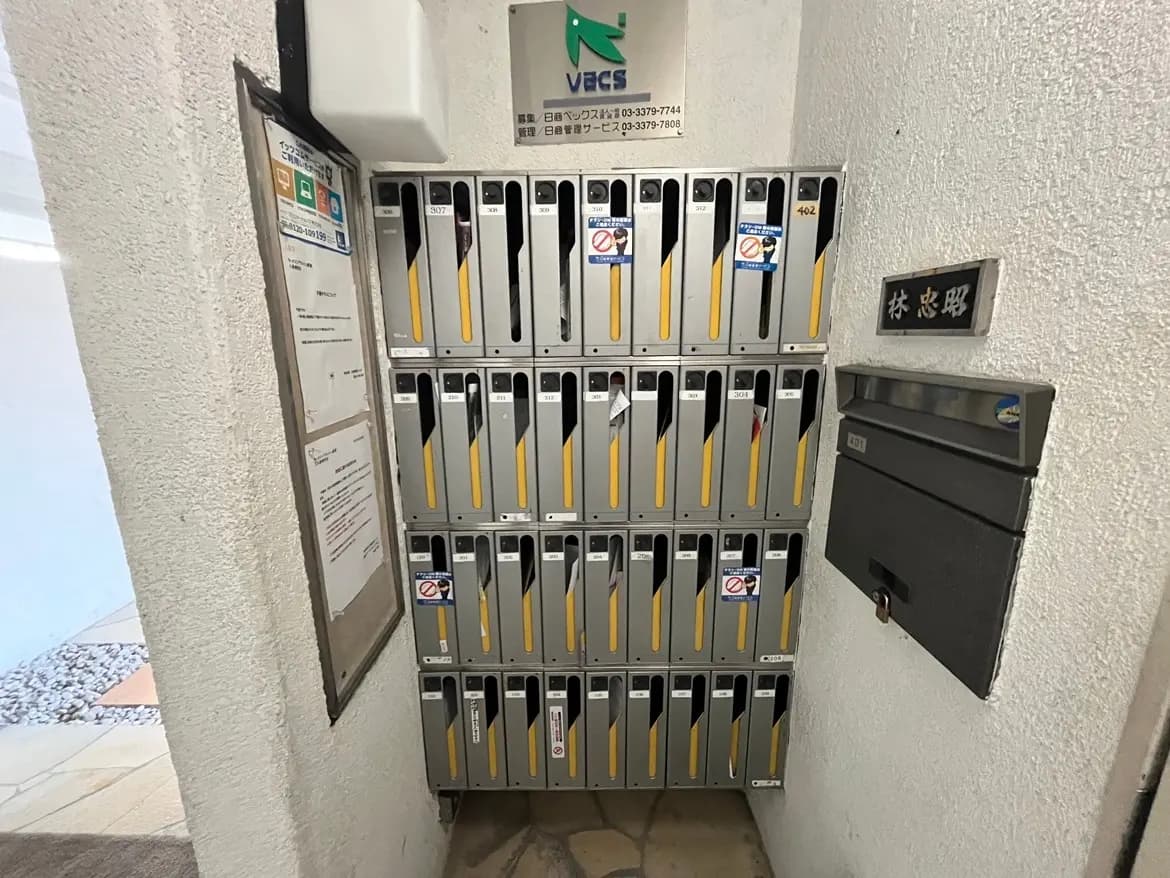 - 住宅のエントランスに配置された郵便受け、番号が付いたロッカー式のメールボックスと掲示板が並ぶ風景