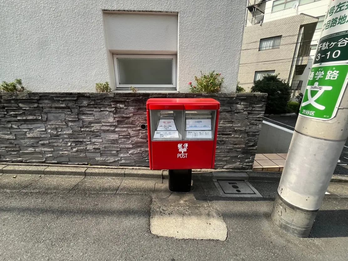 - 街中の赤い郵便ポスト、周囲には石組の壁と植栽があり、近くには通学路の標識が立っている風景