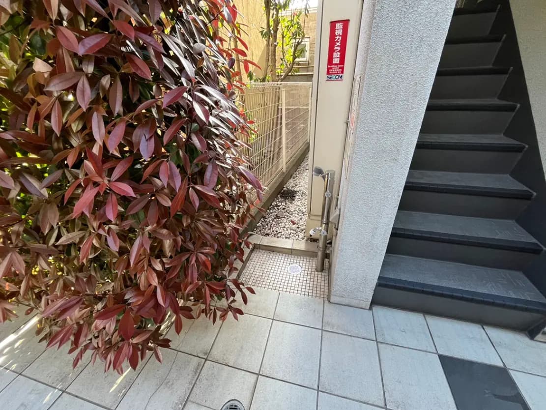 - 赤い葉が印象的な植栽のあるアプローチ、隣接する階段と壁際に設置された監視カメラの設置場所