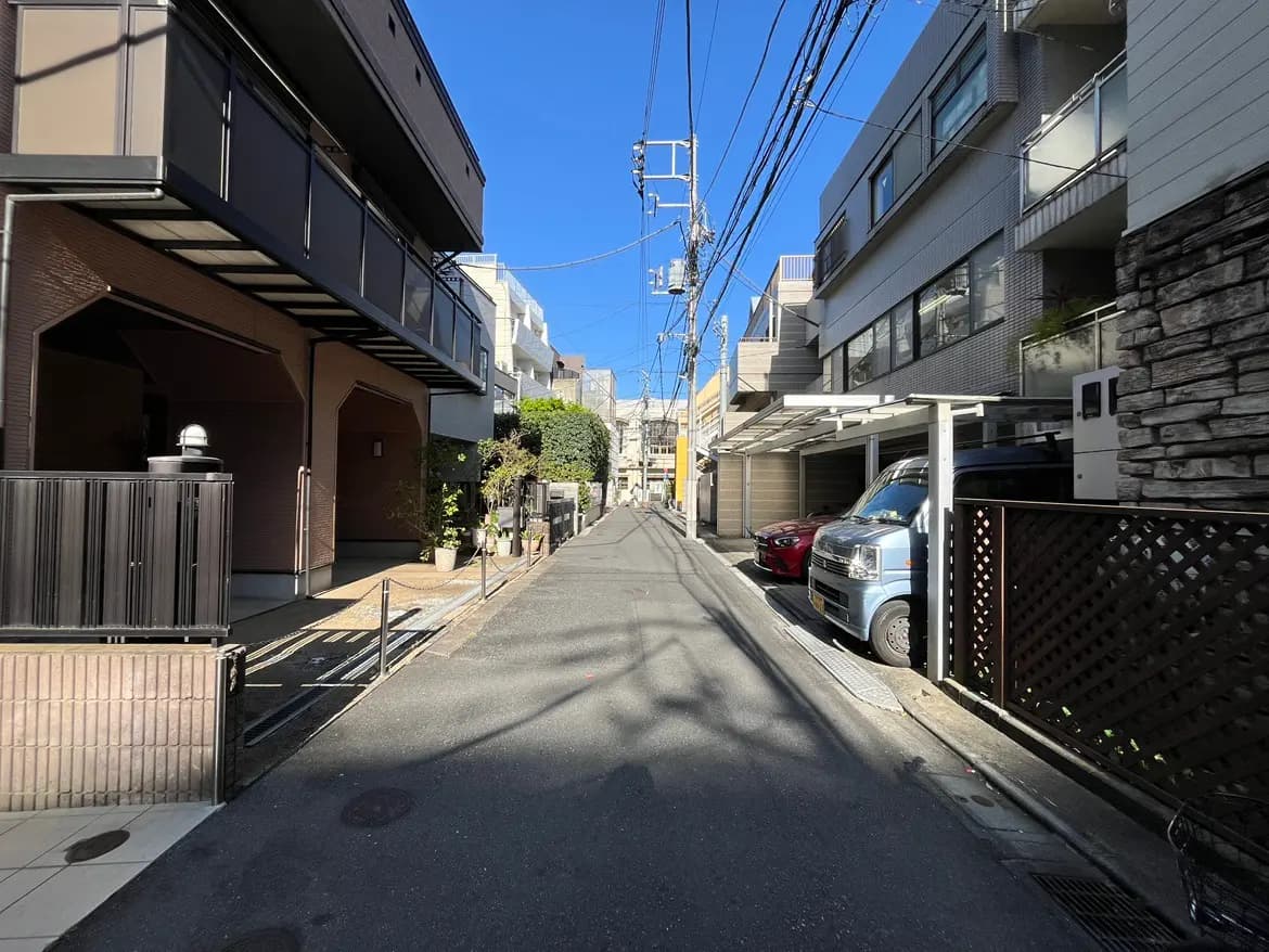 - 明るい青空の下、住宅街の静かな通り、左側にはモダンな建物と植栽、右側には駐車スペースの車が並ぶ風景