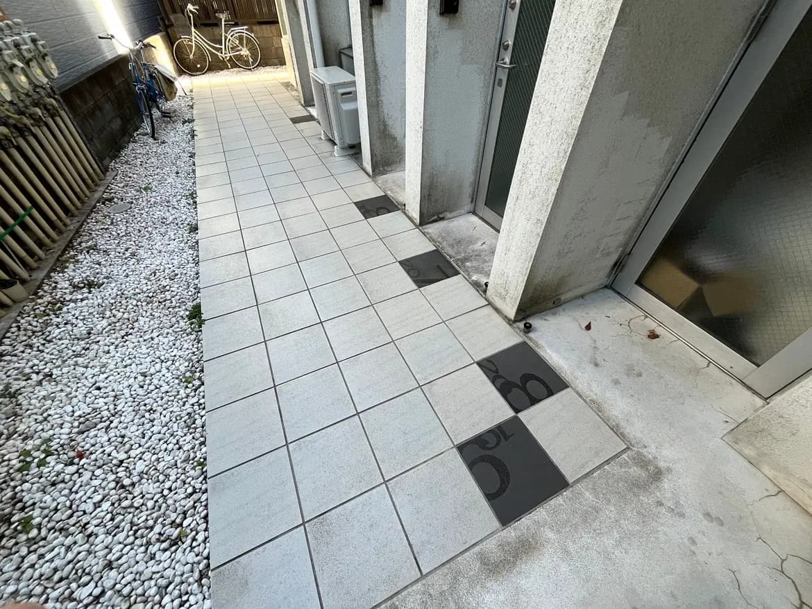 - タイル張りの通路と隣接する白い小石の庭、奥に自転車が見える住宅のエントランス周辺の風景